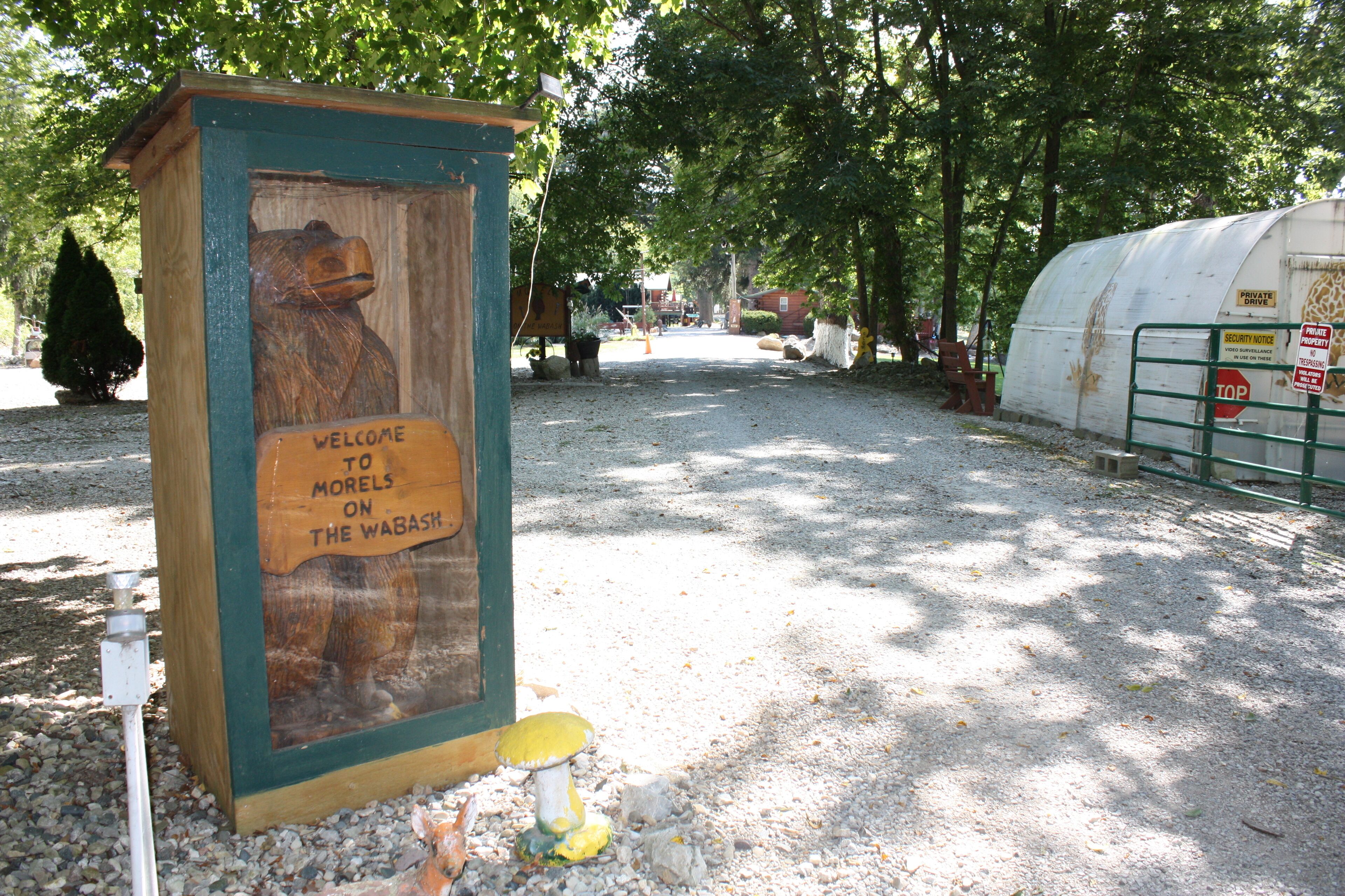 A wooden sign with a bear and a welcome message, a gravel path, trees, and a covered structure in the background.