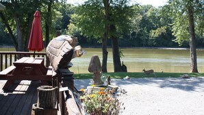 Terraza de madera con mesas de picnic y un paraguas rojo, con vista a un lago donde se ven patos y un ciervo.