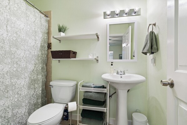 A bathroom with a toilet, sink, and shower curtain. There is a shelf with a plant and a basket, and a towel rack with a towel.