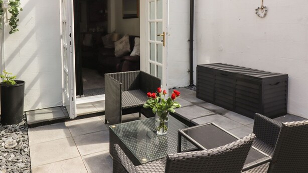 Een terras buiten met rieten meubels, een glazen tafel en een vaas met rode bloemen.