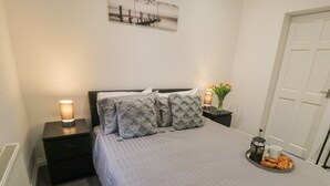 Een netjes opgemaakt bed met een grijze deken en gekleurde kussens, een nachtkastje met een lamp en een bloemenvaas, en een dienblad met gebak.