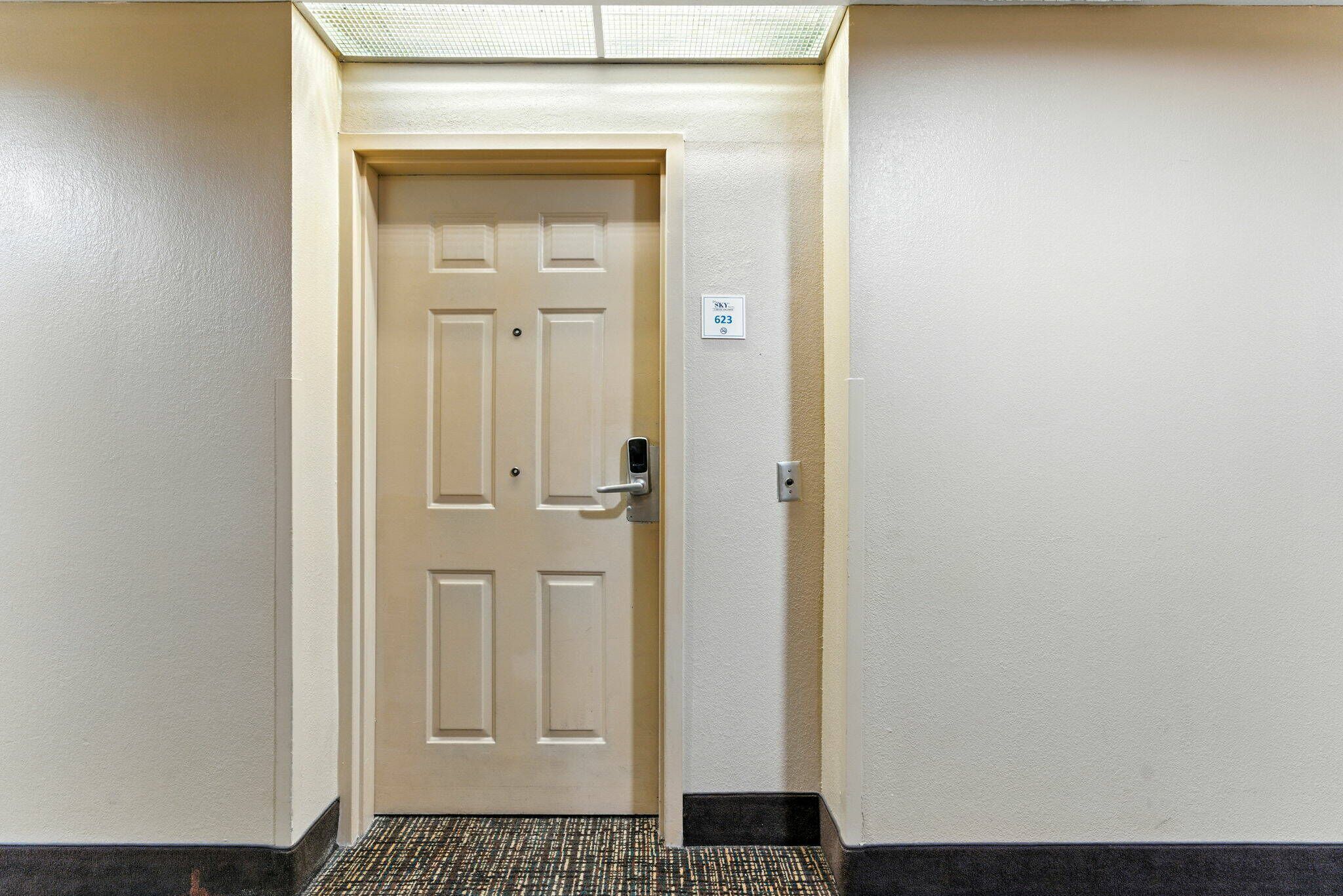 Uma porta bege com fechadura eletrônica, uma pequena placa com o número 623 e um tapete texturizado que leva à porta.