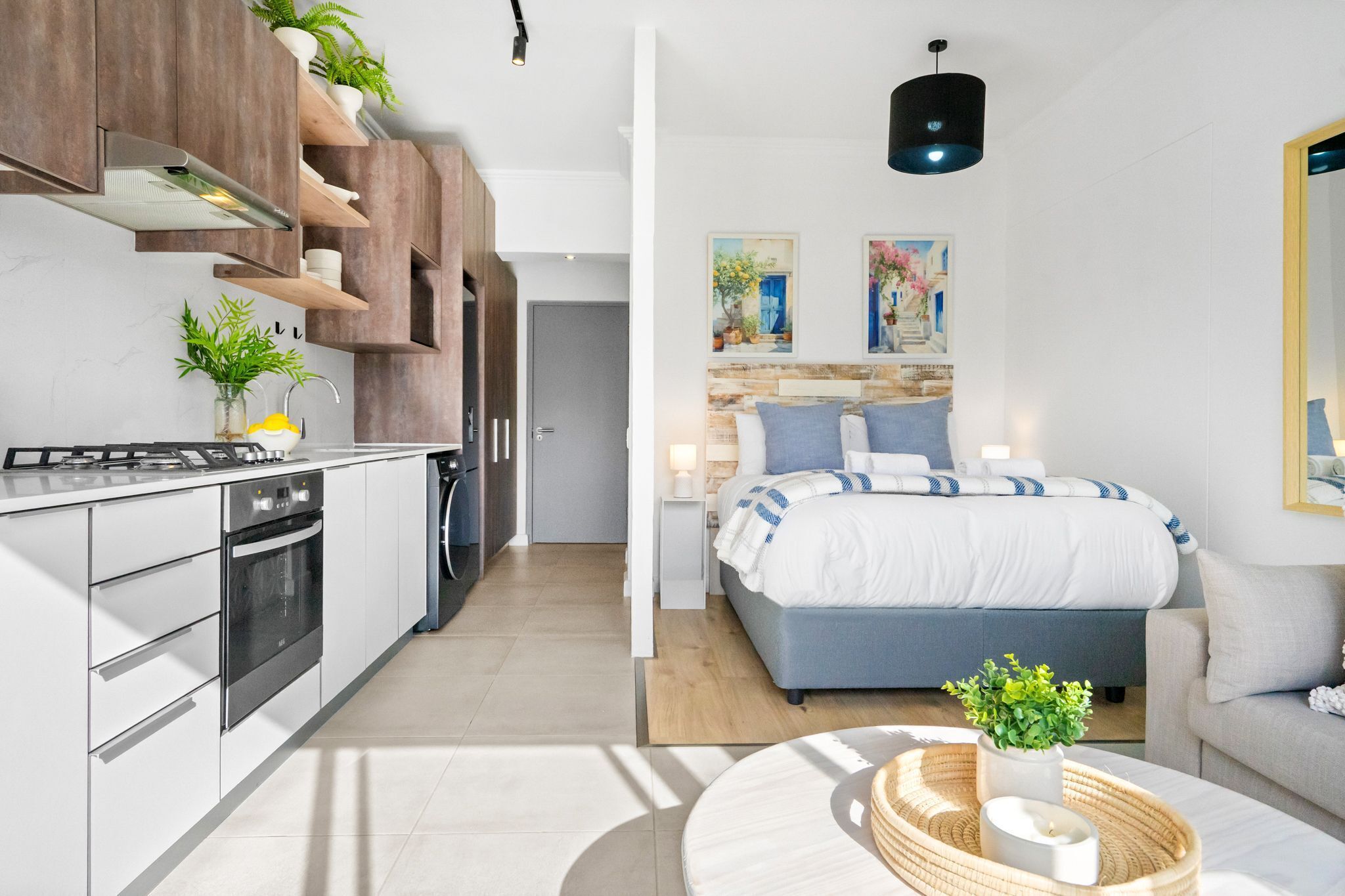 Una cuina moderna amb electrodomèstics integrats, una zona de menjador amb una taula rodona, i un dormitori amb un llit gran i una tauleta de nit.