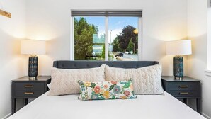 A bedroom with a bed, two bedside tables, two lamps, and a window with a view of the street and trees. - Covelet Hideaway (Blaine)