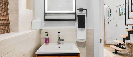 A modern bathroom with a white vanity, a mirror with a light, a towel dispenser, and a wooden cabinet.