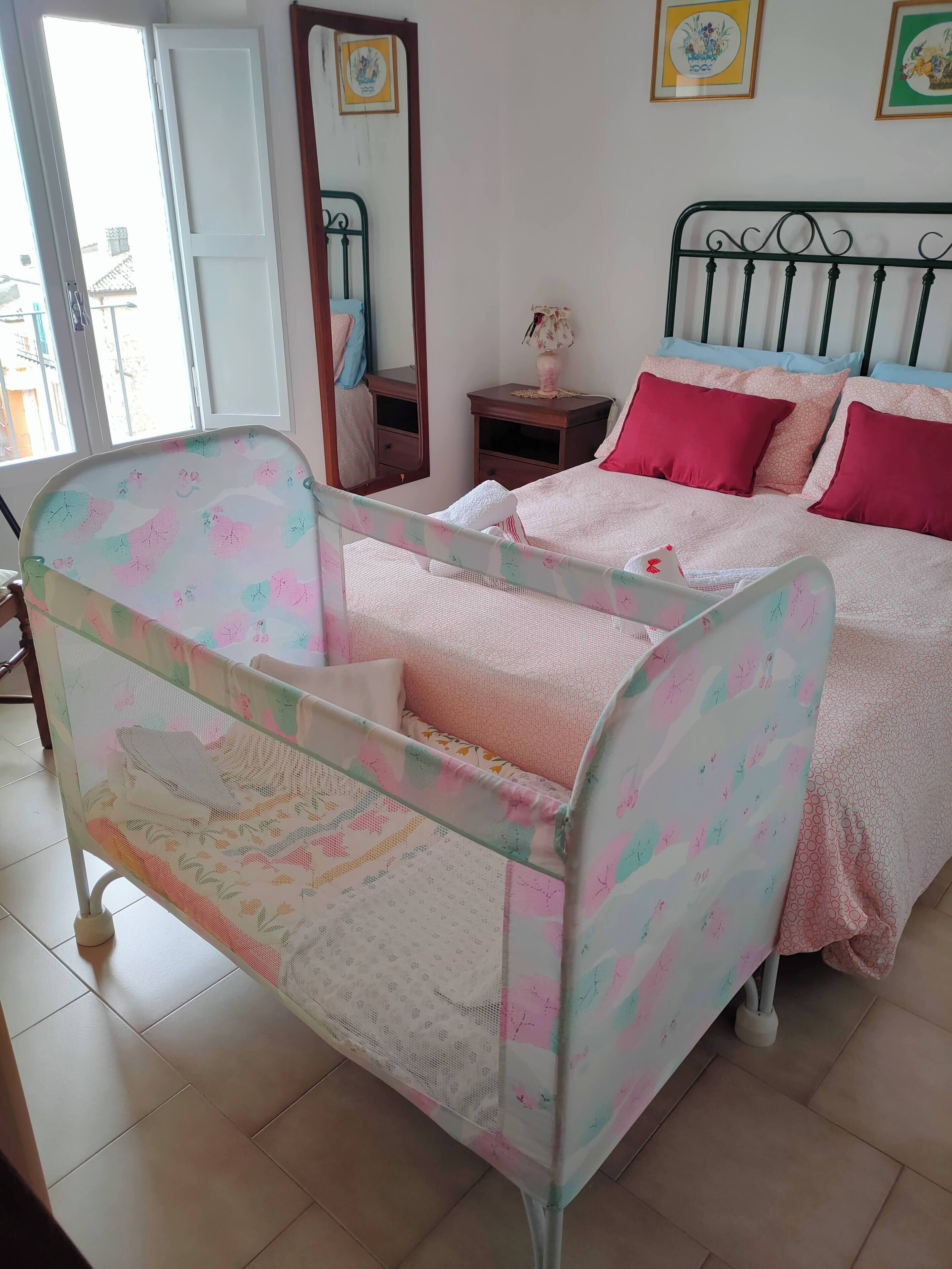 Ein Schlafzimmer mit einem Babybett, einem Bett mit rosa Bettwäsche, einem Nachttisch und einem Spiegel.