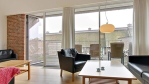 Ein modernes Wohnzimmer mit einer großen Schiebeglaswand, die auf einen Balkon führt, einem schwarzen Ledersofa, einem Holztisch und einer hängenden Pendelleuchte.