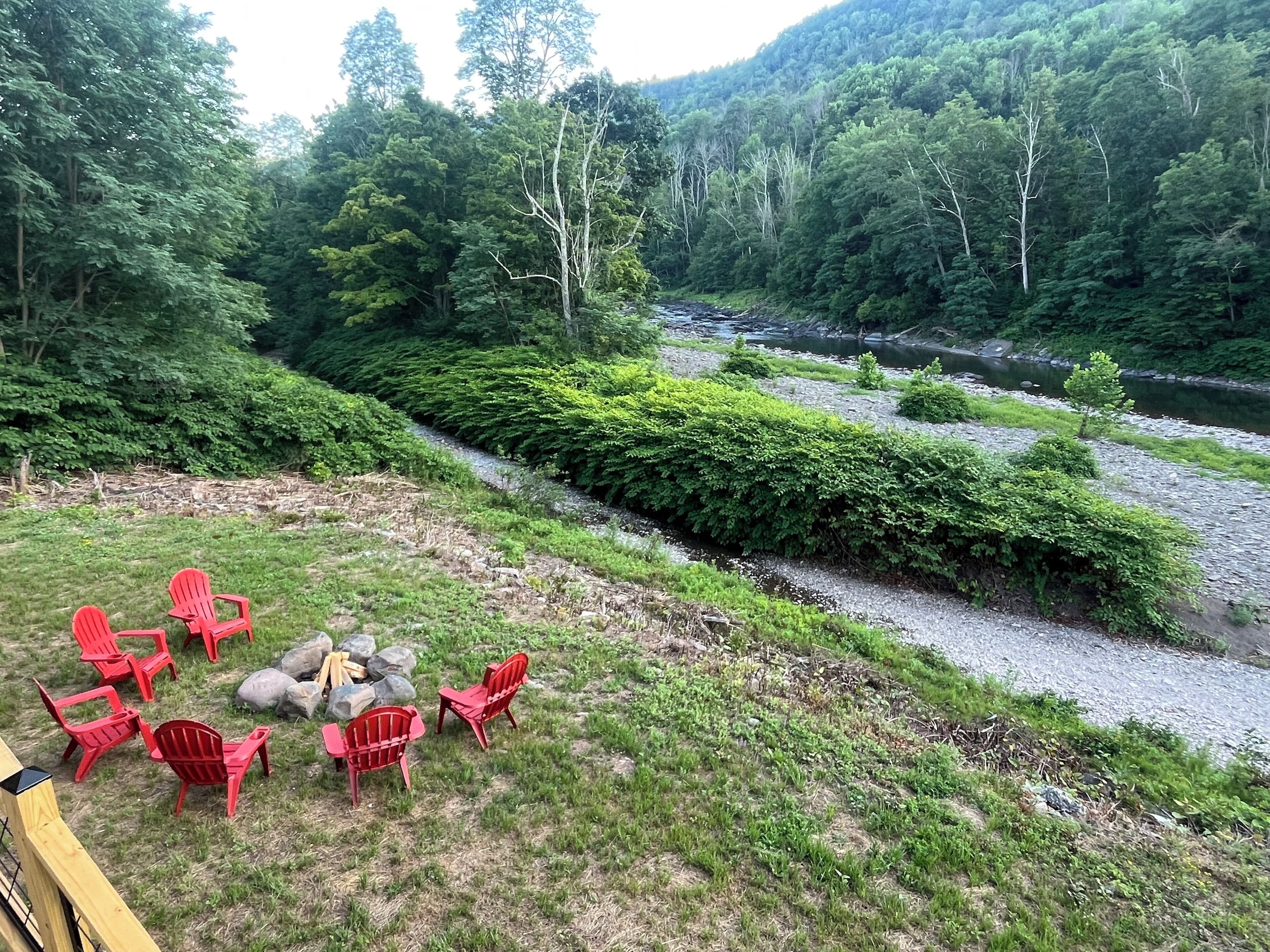 Un espacio con asientos a la orilla del río, con sillas rojas y una chimenea, rodeado de vegetación exuberante y una ladera boscosa.