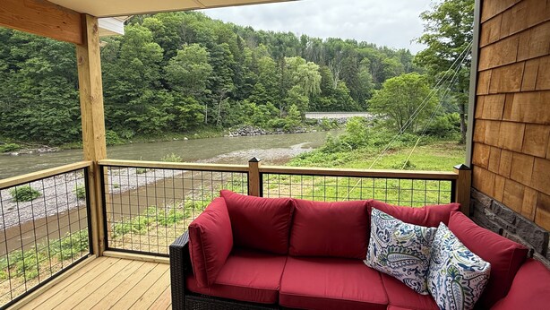 Een rode bank met gekleurde kussens op een houten terras met uitzicht op een rivier en bos.