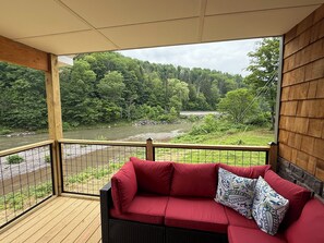 A red sofa with patterned cushions on a wooden deck overlooking a river and forest. - Charming 3-bedroom house with WiFi, AC in Historic Prattsville. (Prattsville)