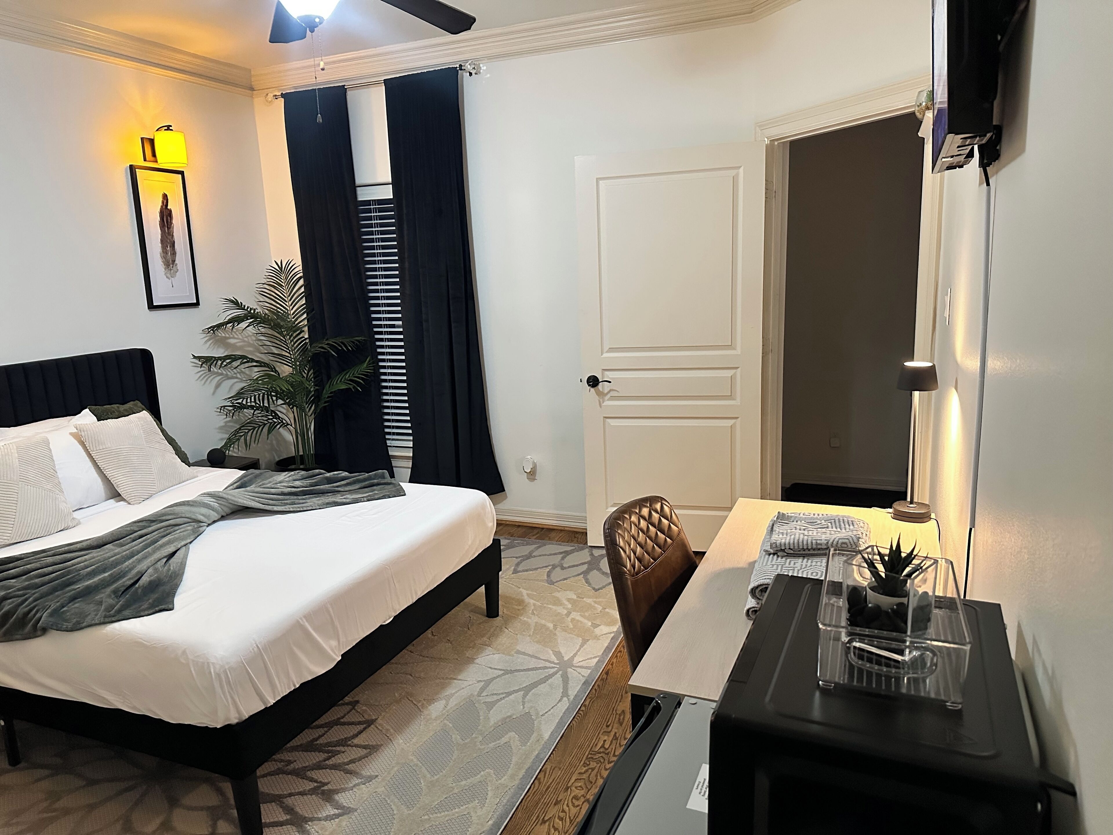 Habitación de hotel ordenada con cama, escritorio, silla, planta en maceta y puerta.