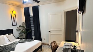 Ein ordentlich eingerichtetes Hotelzimmer mit einem Bett, einem Schreibtisch, einem Stuhl, einer Topfpflanze und einer Tür.