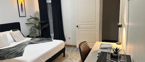 Habitación de hotel ordenada con cama, escritorio, silla, planta en maceta y puerta.