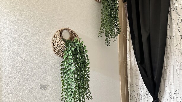 Ein Zimmer mit strukturierter Wand, zwei gewebten Wandbehängen mit hängenden Pflanzen und einem Vorhang mit Muster.