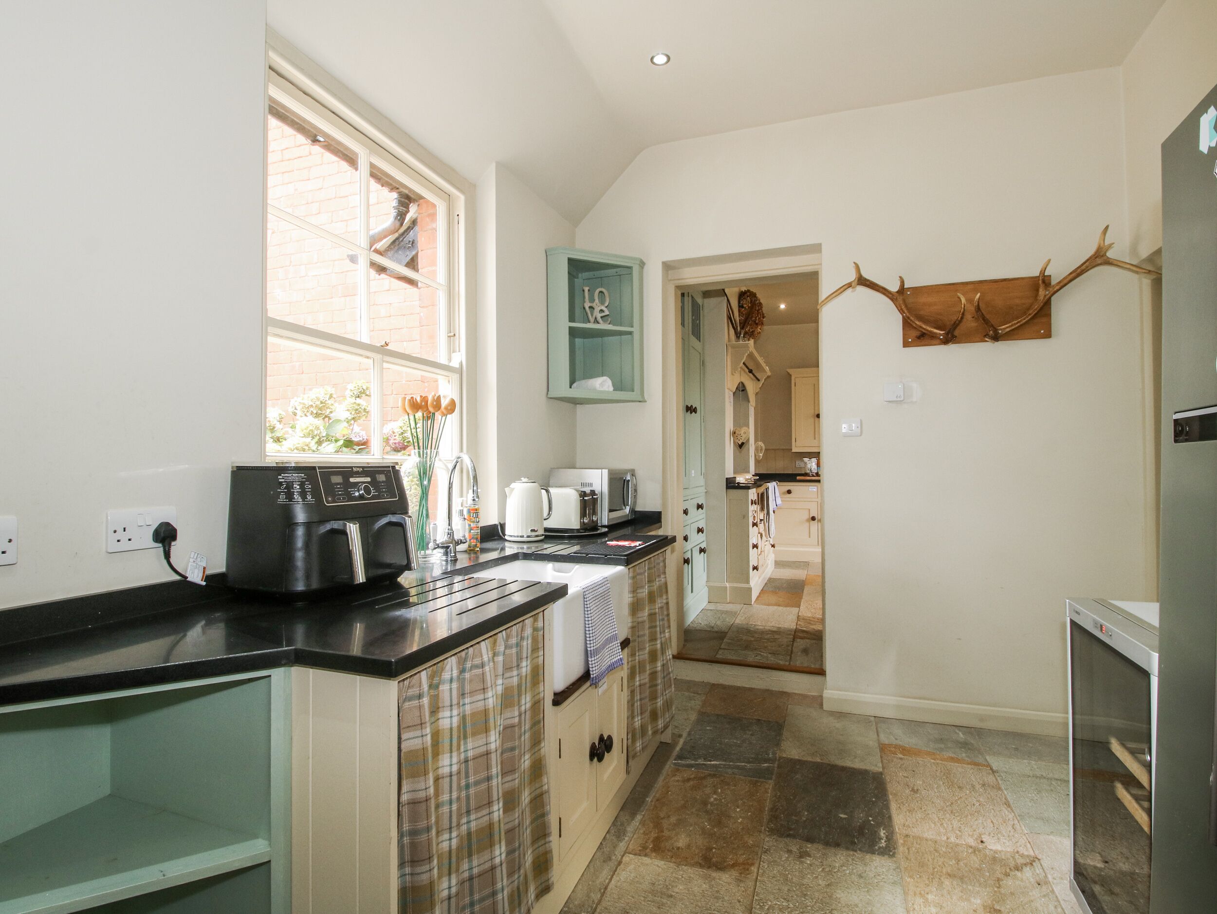 A kitchen with a coffee machine, a kettle, and a toaster. There is a window with a view of a brick wall and flowers. A wooden antler decoration hangs on the wall.