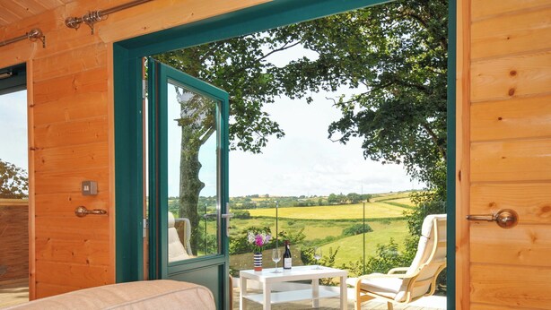 A wooden room with a glass door leading to an outdoor seating area with a table, chairs, and a view of a green landscape. - Owl Lodge (Liskeard)