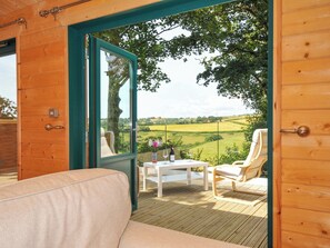 A wooden room with a glass door leading to an outdoor seating area with a table, chairs, and a view of a green landscape. - (Liskeard)