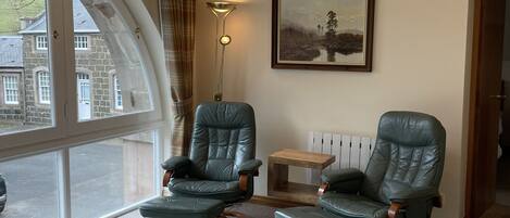 A room with two leather armchairs, a small wooden table, and a sofa. A large window offers a view of a stone building outside.