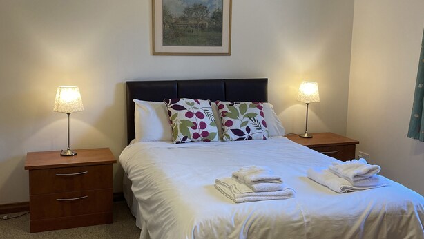 Una cama bien hecha con ropa blanca y almohadones decorativos, flanqueada por dos mesitas de noche con lámparas, en una habitación sencilla con un cuadro en la pared.