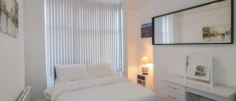 A bedroom with a bed, a bedside table with a lamp, a dresser, and a large window with blinds.