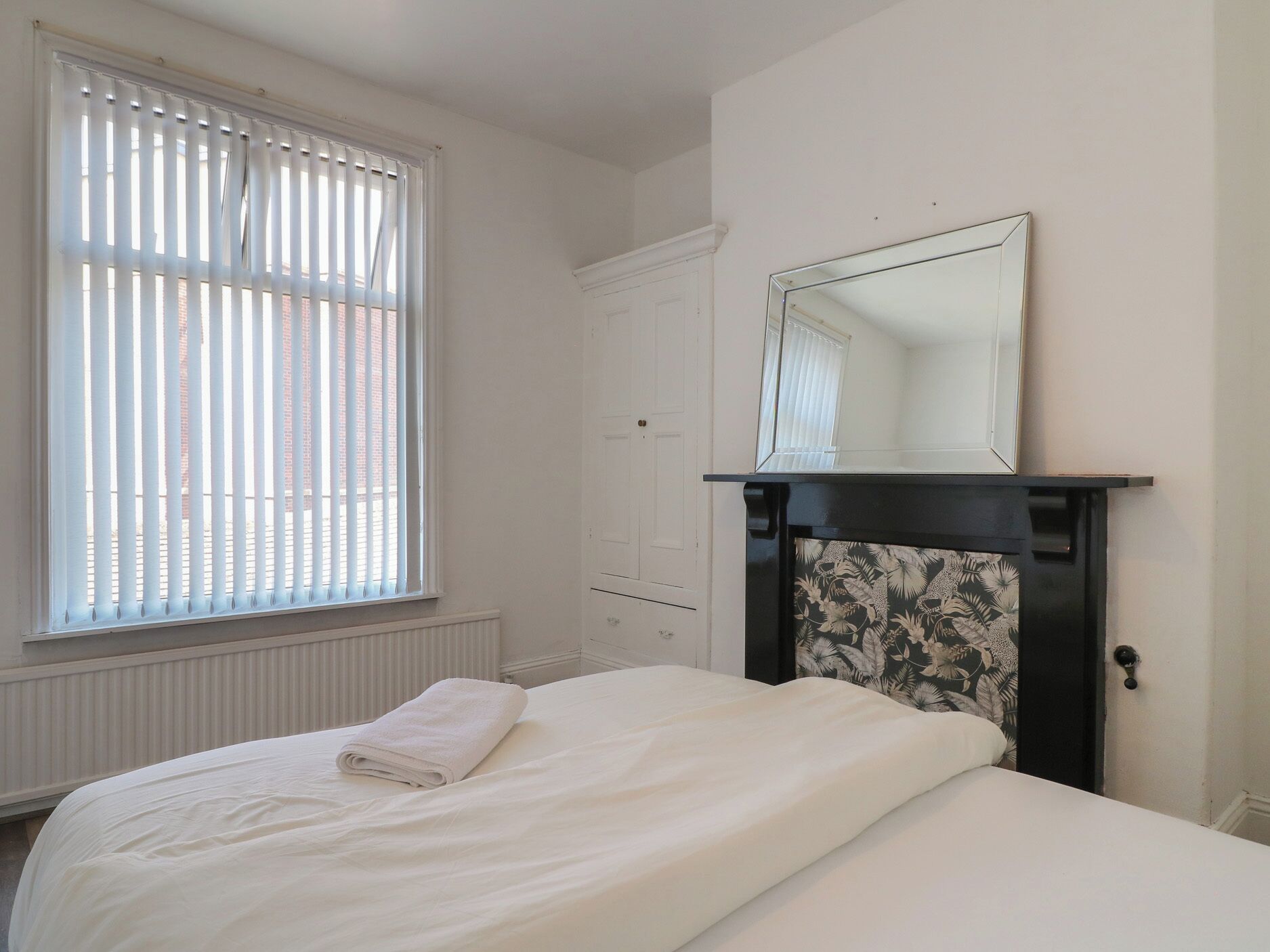 A bedroom with a bed, a window with blinds, a door, and a fireplace with a mirror above it.