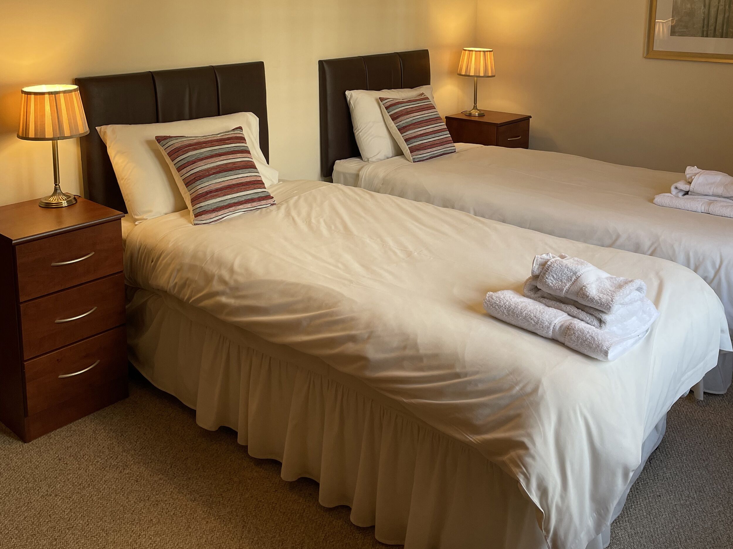 Two neatly made beds with white linens, striped pillows, and matching nightstands with lamps.