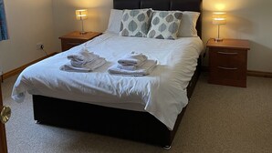 Cama bem arrumada com lençóis brancos e duas toalhas, acompanhada por duas mesas de cabeceira com abajures, num quarto com design simples e limpo.