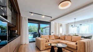 A modern living room with a beige sectional sofa, a coffee machine, and a dining area with a table and chairs. - Charming 3BR Villa with Heated Pool, Sauna and Gym (Privlaka)