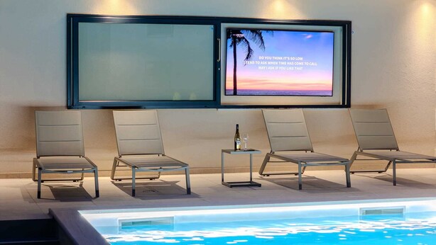 A poolside area with four lounge chairs, a small table with a bottle and glass, and a large screen displaying a sunset scene. - Charming 3BR Villa with Heated Pool, Sauna and Gym (Privlaka)