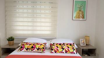 A hotel room with a bed, colorful pillows, a framed pineapple picture, a small plant, and a remote control on a nightstand.