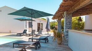 A wooden deck with lounge chairs and umbrellas, a pool, and a building with a tiled roof. - Villa Meia Di Porto-Vecchio (PORTO VECCHIO)