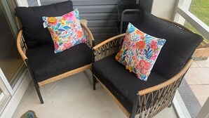 Two wicker chairs with black cushions and colorful floral pillows on a porch.