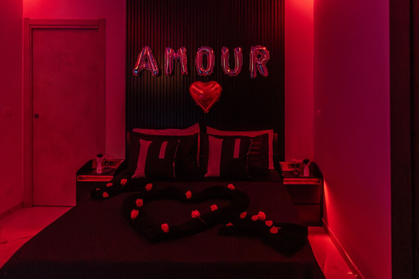 Ein schwach beleuchteter Raum mit einem Bett, einem Nachttisch und einer Wand mit der Aufschrift 'AMOUR' und einem Herzsymbol.