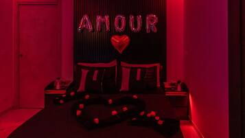 Ein schwach beleuchteter Raum mit einem Bett, einem Nachttisch und einer Wand mit der Aufschrift 'AMOUR' und einem Herzsymbol.