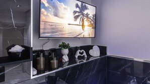 A modern bathroom with a large TV mounted above a black marble countertop, a potted plant, and decorative heart-shaped objects. - Amour - House, Private Jacuzzi (Rosolini)