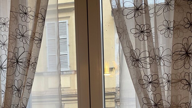 A window with sheer curtains featuring a floral pattern, a white door, and a view of a building with balconies and flowers outside. - Chagall Apartment - no ZTL (Asti)