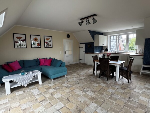 A cozy living room with a teal sofa, a dining table with chairs, and a kitchen area with white cabinets and a window.