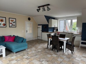 A cozy living room with a blue sofa, a dining area with wicker chairs, a kitchen with white cabinets, and a large window.