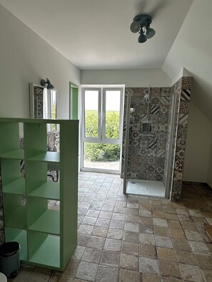 A bathroom with a shower, a green shelving unit, and a view of greenery outside.
