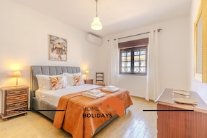 Une chambre à coucher comprenant un lit, des tables de chevet, une fenêtre avec des rideaux et un climatiseur fixé au mur.