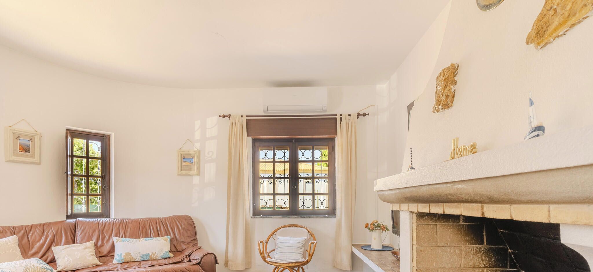 Een gezellige woonkamer met een bruine leren bank, een houten bijzettafeltje en rieten stoelen. Er is een open haard met een bakstenen omlijsting en een raam met gordijnen.