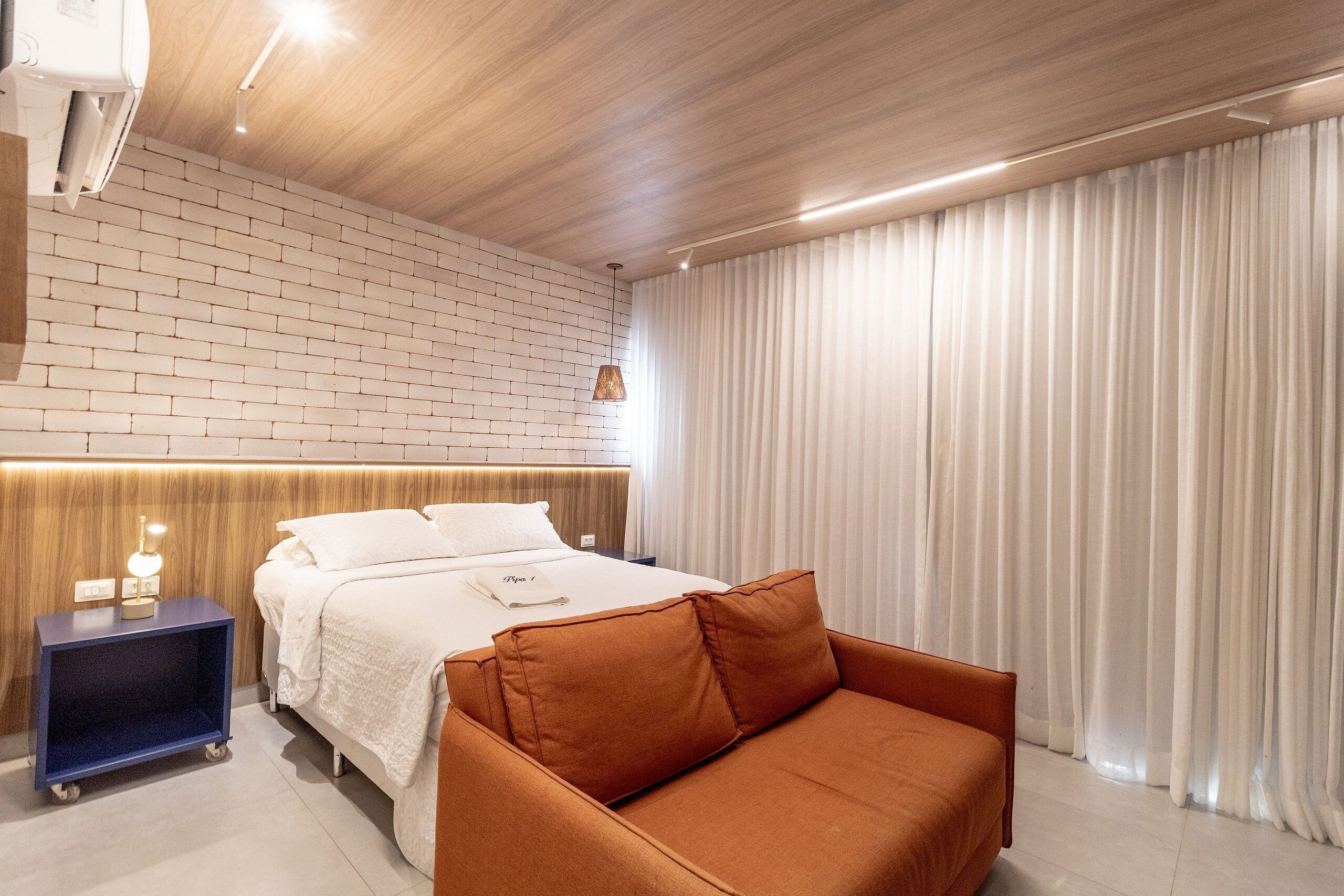 Une chambre d’hôtel moderne avec un mur au motif de briques, un plafond en bois, un lit avec une literie blanche, un canapé marron et une grande fenêtre avec des rideaux blancs.