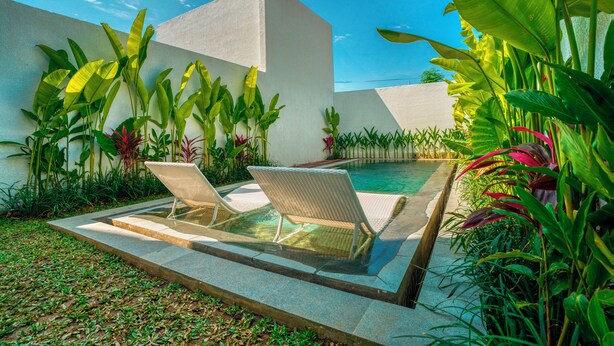 A poolside area with two lounge chairs, surrounded by lush tropical plants and a white building.
