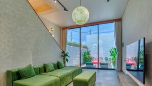 A modern living room with a green sofa, a round pendant light, and a large window overlooking a balcony with plants and a pink flamingo statue.
