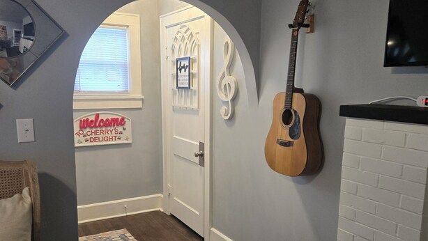 A room with a wooden guitar, a TV, a white brick fireplace, and a door leading to another room with a sign. - 🍒CHERRY’S DELIGHT - Where timeless comfort is served with a cherry on top! (Glasgow)