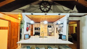A kitchen with wooden cabinets, a central island with bar stools, hanging lights, and a ship wheel on the ceiling. - Beachside Get-A-Way! Walk to the Beach! (Galveston)