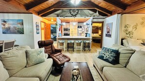 A cozy living room with a high ceiling, wooden beams, and a central island with bar stools. There are two sofas and a coffee table with decorative items. - Beachside Get-A-Way! Walk to the Beach! (Galveston)