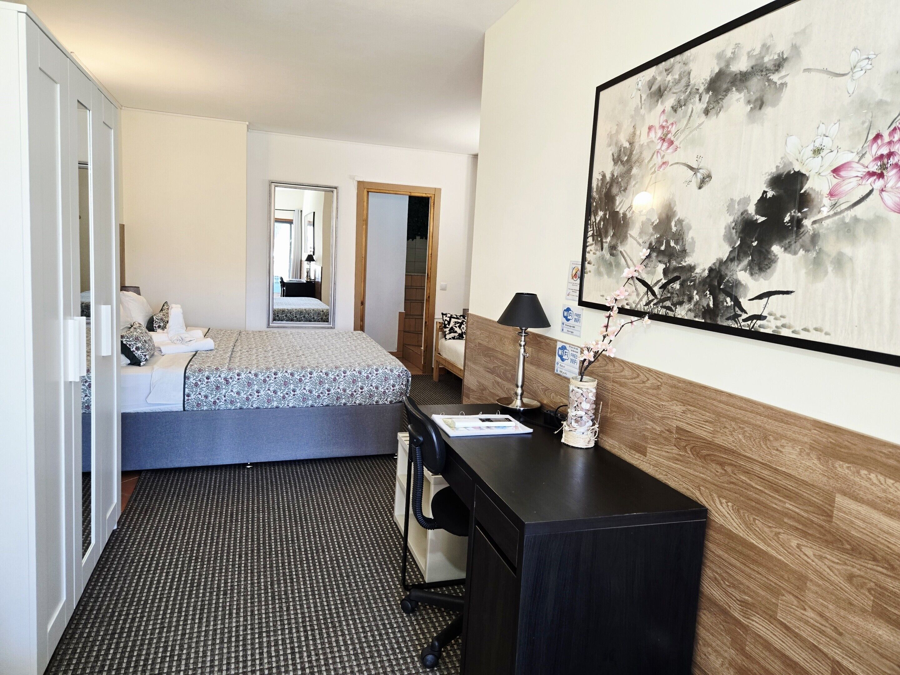 Hotellihuone, jossa on sänky, työpöytä, tuoli, lamppu, maljakko kukkineen ja taulu seinällä.