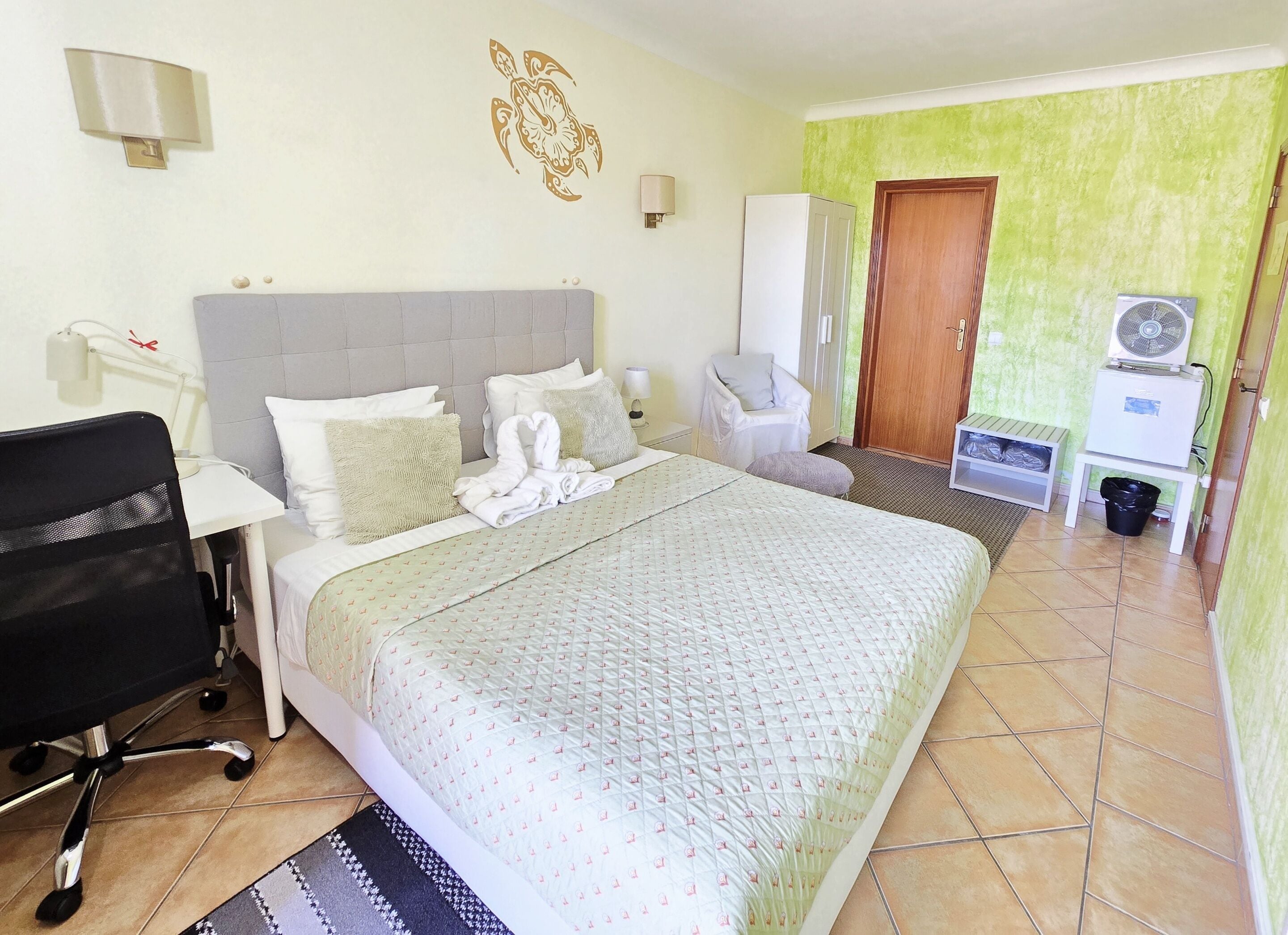 Une chambre d’hôtel avec un grand lit, un bureau, une chaise, une armoire, un ventilateur et une poubelle.