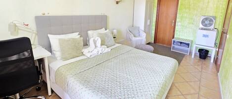 Une chambre d’hôtel avec un grand lit, un bureau, une chaise, une armoire, un ventilateur et une poubelle.